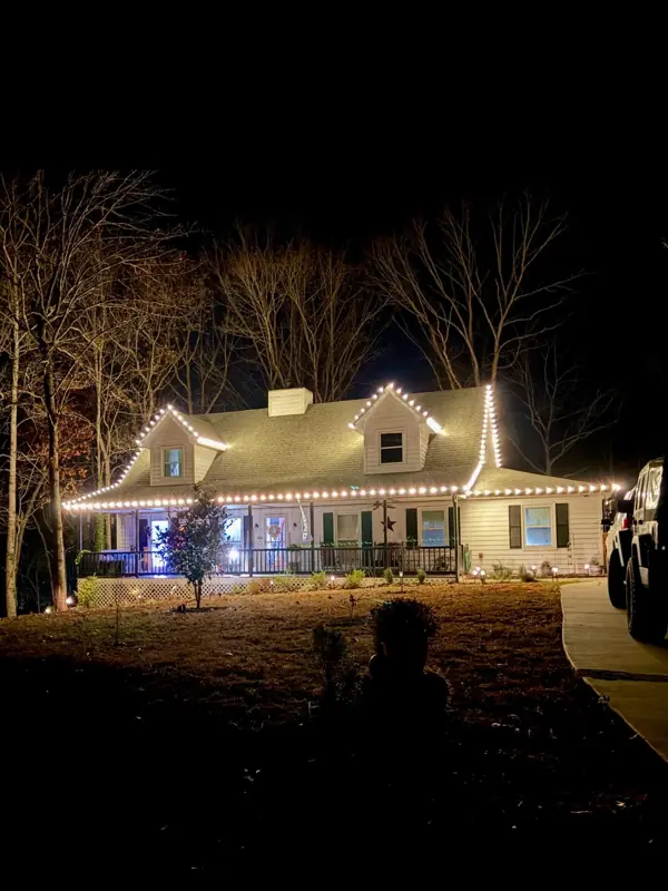 Cape Cod style home among tall trees at night with warm white lights outlining the roofline and dormers