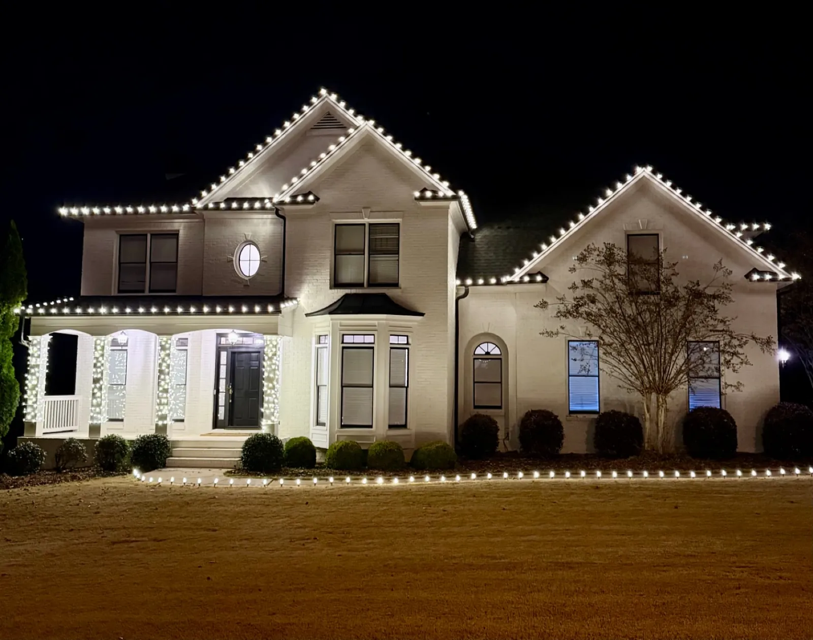 White two-story brick home at night with warm white lights along every roofline and peak, wrapped porch columns, and a lit walkway
